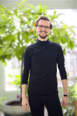 Max Frühschütz vor einem unscharfen Pflanzenhintergrund. Er trägt eine Hornbrille, ein schwarzes Shirt, hat einen gepflegten Drei-Tage-Bart und lächelt in die Kamera